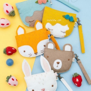 May include: A collection of felt animal-themed coin purses in various colors, including yellow, orange, blue, brown, and white. Each purse features a different animal face, such as a fox, bear, and bunny. The purses have wrist straps and the text "iThinkSew.com".