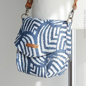Puede incluir: Un bolso bandolera azul y blanco con estampado, con una correa de cuero marrón y un cierre de metal. El bolso tiene una solapa con una etiqueta de cuero marrón que dice "Handmade".