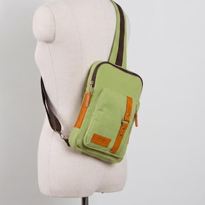 May include: A light green sling bag with dark brown trim and a single shoulder strap. The bag features a front pocket with a vertical tan leather-like accent strap, a small rectangular patch with text, and dark brown zippers with tan pull tabs.