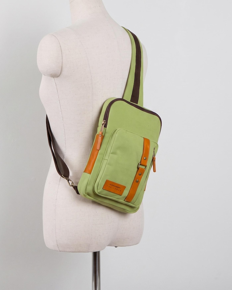 May include: A light green sling bag with dark brown trim and a single shoulder strap. The bag features a front pocket with a vertical tan leather-like accent strap, a small rectangular patch with text, and dark brown zippers with tan pull tabs.