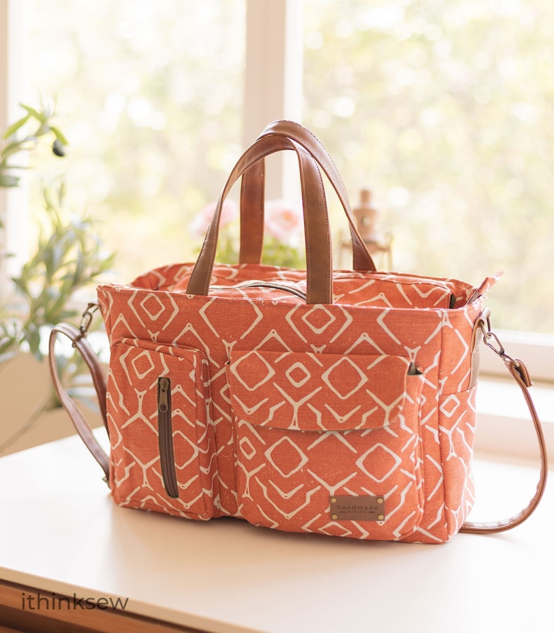 K&ouml;nnte beinhalten: Eine gro&szlig;e orangefarbene Canvas-Tragetasche mit geometrischem Muster und braunen Ledergriffen. Die Tasche hat eine Rei&szlig;verschlusstasche auf der Vorderseite und ein Etikett mit der Aufschrift "Handmade with Love".