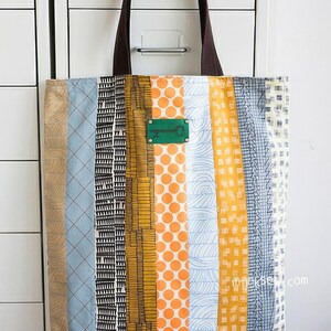 K&ouml;nnte beinhalten: Eine gestreifte Tragetasche mit verschiedenen Mustern in Blau, Orange, Gelb und Schwarz. Die Tasche hat braune Ledergriffe und ein gr&uuml;nes Metalletikett mit einem Schl&uuml;sselloch-Design.