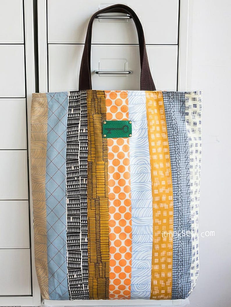 K&ouml;nnte beinhalten: Eine gestreifte Tragetasche mit verschiedenen Mustern in Blau, Orange, Gelb und Schwarz. Die Tasche hat braune Ledergriffe und ein gr&uuml;nes Metalletikett mit einem Schl&uuml;sselloch-Design.