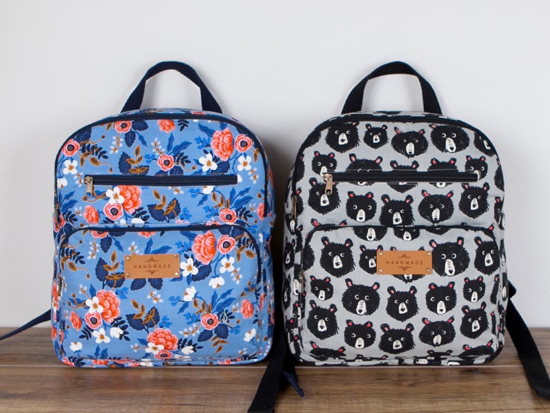 May include: Two handmade backpacks, one with a floral pattern on a light blue background and the other with a black bear pattern on a grey background. Both backpacks have a black zipper and a brown leather tag that says "Handmade".