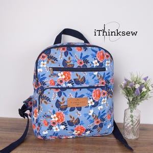 Puede incluir: Una mochila de color azul claro con un estampado floral de flores y hojas rojas, blancas y azul oscuro. La bolsa tiene un asa superior azul oscuro, dos bolsillos delanteros con cremallera y una etiqueta marrón con la palabra HANDMADE.