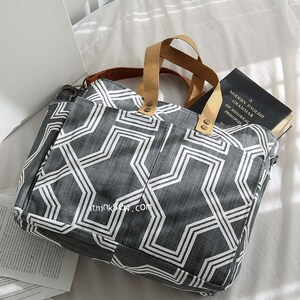 May include: A gray and white geometric patterned tote bag with brown leather handles. The bag is lying on a white surface with a book inside.