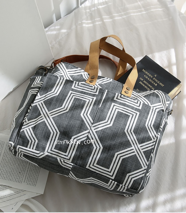 May include: A gray and white geometric patterned tote bag with brown leather handles. The bag is lying on a white surface with a book inside.