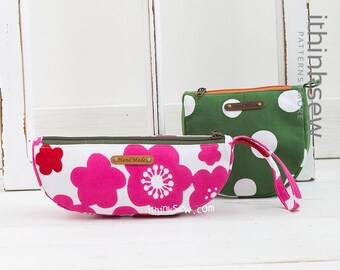 May include: Two fabric zippered pouches with a curved shape. The pouch on the left has a pink and white floral print with a brown "Hand Made" label. The pouch on the right has a green background with white polka dots and a brown "Hand Made" label. Both pouches have a brown zipper and a brown handle.