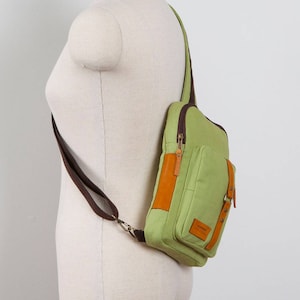 May include: A light green canvas sling bag featuring brown fabric straps, dark brown zippers, and tan leather accents. The bag includes a front pocket with a small rectangular leather patch displaying text, secured by a vertical leather strap and metal studs.