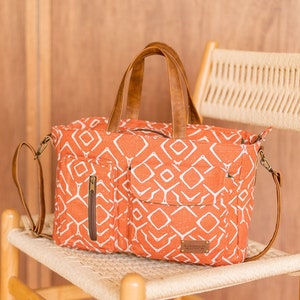 K&ouml;nnte beinhalten: Eine gro&szlig;e orangefarbene Canvas-Tragetasche mit geometrischem Muster und braunen Ledergriffen und -riemen. Die Tasche hat eine Rei&szlig;verschlusstasche auf der Vorderseite und ein Etikett mit der Aufschrift "Handmade".