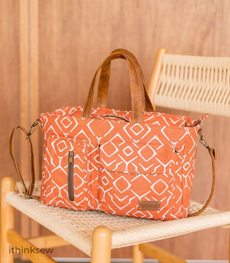 K&ouml;nnte beinhalten: Eine gro&szlig;e orangefarbene Canvas-Tragetasche mit geometrischem Muster und braunen Ledergriffen und -riemen. Die Tasche hat eine Rei&szlig;verschlusstasche auf der Vorderseite und ein Etikett mit der Aufschrift "Handmade".