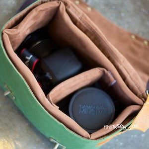 Pu&ograve; includere: Borsa per fotocamera in tela verde con dettagli in pelle marrone e interno in tessuto marrone. La borsa &egrave; aperta e contiene un obiettivo per fotocamera nero e un tappo per obiettivo per fotocamera nero con la scritta "SAMSUNG MINOLTA" stampata su di esso.