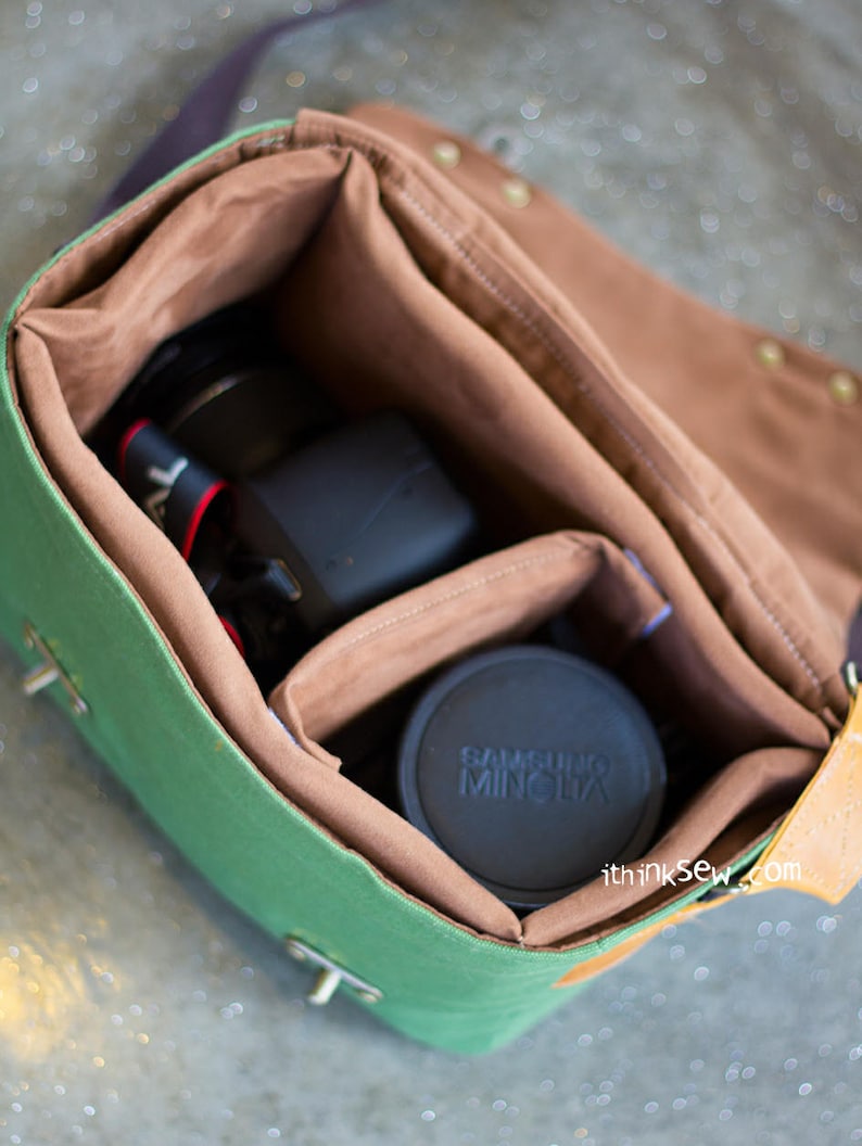 Pu&ograve; includere: Borsa per fotocamera in tela verde con dettagli in pelle marrone e interno in tessuto marrone. La borsa &egrave; aperta e contiene un obiettivo per fotocamera nero e un tappo per obiettivo per fotocamera nero con la scritta "SAMSUNG MINOLTA" stampata su di esso.