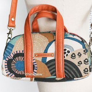May include: A small, brown leather handbag with a crossbody strap. The bag has a geometric pattern with circles, lines, and flowers in shades of blue, brown, and pink. The bag has a metal clasp and a "HANDMADE" label.