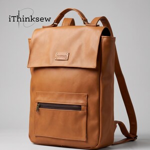 Peut inclure: Un sac à dos marron clair avec un rabat supérieur, une poignée et des bretelles réglables. L'avant présente une poche zippée et une étiquette rectangulaire avec le mot handmade. Le texte iThinksew est visible dans le coin supérieur gauche.
