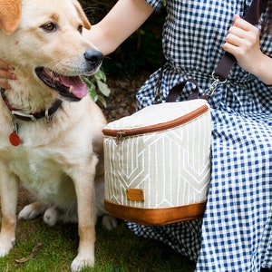 May include: A beige and brown insulated lunch bag with a geometric pattern and a brown leather trim. The bag is being carried by a person wearing a blue and white gingham dress. A dog is sitting next to the person.