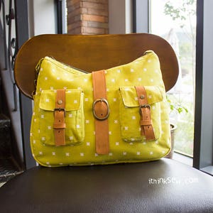 May include: A yellow shoulder bag with white floral patterns and two front pockets. The bag features brown leather straps and buckles, and a large brown leather strap with a buckle closure. The bag is sitting on a wooden chair.