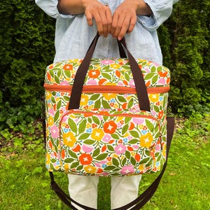 May include: A floral cooler bag with brown straps and a shoulder strap. The bag has a pattern of orange, yellow, pink, and blue flowers with green leaves on a white background. It features an orange zip and a front pocket. The bag is designed for carrying items.