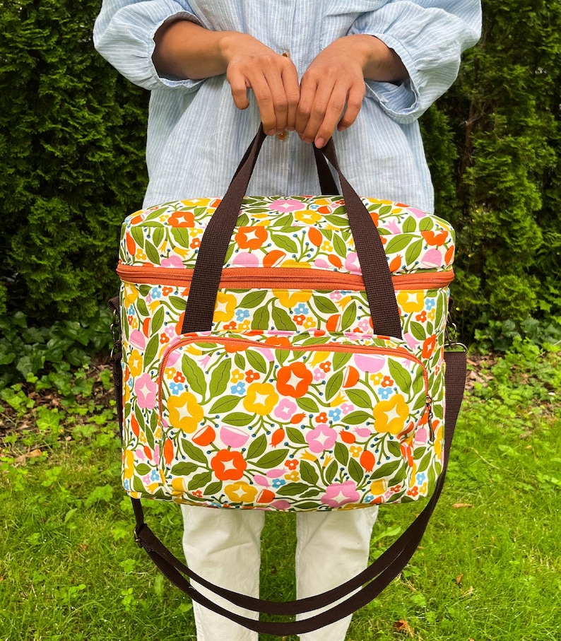 May include: A floral cooler bag with brown straps and a shoulder strap. The bag has a pattern of orange, yellow, pink, and blue flowers with green leaves on a white background. It features an orange zip and a front pocket. The bag is designed for carrying items.