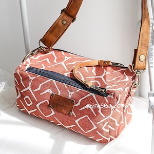 May include: A small, red and white patterned canvas bag with a brown leather strap and a zipper closure. The bag has a rectangular shape and is perfect for carrying small items.