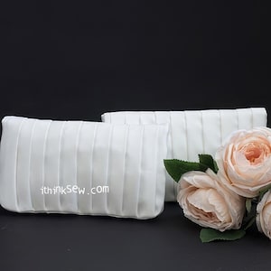 Puede incluir: Dos bolsos de mano blancos de satén con detalles plisados. Los bolsos están sobre una superficie negra con un ramo de rosas color melocotón.