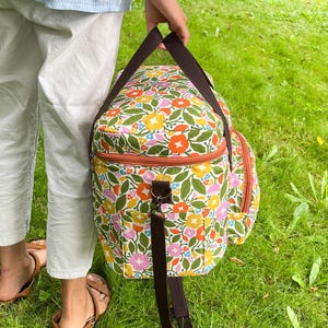 May include: A colourful floral bag with brown straps and a zip. The bag has a pattern of orange, yellow, pink, and blue flowers on a white background. The bag is being held by a person on a grassy lawn.