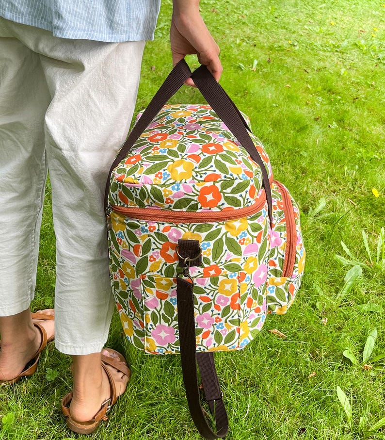 May include: A colourful floral bag with brown straps and a zip. The bag has a pattern of orange, yellow, pink, and blue flowers on a white background. The bag is being held by a person on a grassy lawn.