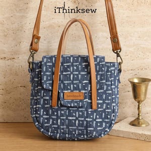Pode incluir: Uma bolsa de tecido com padrão azul e branco, alças de couro castanho-claro e uma alça de ombro removível. A bolsa possui um fecho de aba com um pequeno remendo retangular de couro castanho-claro com a palavra handmade. O texto iThinksew é visível.