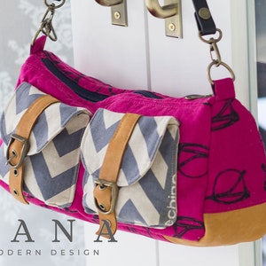 May include: A pink and grey canvas shoulder bag with a brown leather strap and two front pockets. The bag has a chevron pattern on the front pockets and a black and white print on the main body. The bag is labelled "DANA MODERN DESIGN".