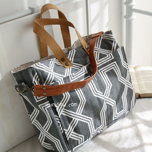 May include: A large gray and white geometric patterned tote bag with brown leather straps and handles. The bag has a zipper closure and a small pocket on the front. The bag is sitting on a white surface.