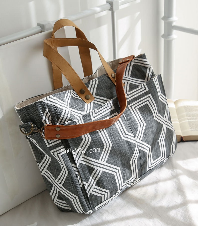 May include: A large gray and white geometric patterned tote bag with brown leather straps and handles. The bag has a zipper closure and a small pocket on the front. The bag is sitting on a white surface.