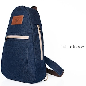 Pode incluir: Uma mochila de ganga azul escura com um remendo de couro castanho e um bolso com fecho de correr. A mochila tem uma única correia e um pequeno bolso na frente.