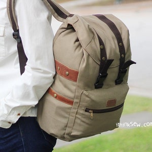 May include: A tan canvas backpack with brown leather accents and buckles. The backpack has a large main compartment and a smaller front pocket with a zipper. The backpack is worn by a person wearing a white shirt and blue jeans.