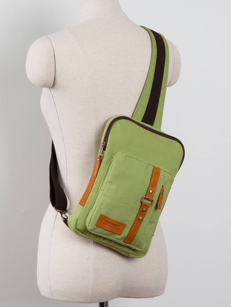 May include: A light green canvas sling bag featuring dark brown trim, a single wide shoulder strap, and a front pocket secured with a brown leather-like strap and metal snap buttons. A small rectangular leather-like patch is visible on the front.