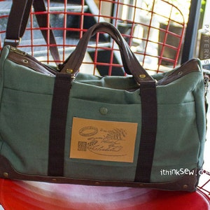 May include: A green canvas duffel bag with brown leather trim and a brown leather handle. The bag has a brown leather patch with a design and the text "i think sew" on it.
