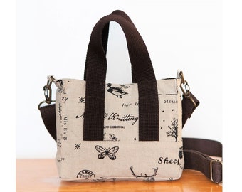May include: A small beige tote bag with brown handles and a matching adjustable shoulder strap. The bag features a repeating pattern of black illustrations and text, including a butterfly, a bird, and the words "Knitting and Embroidery."