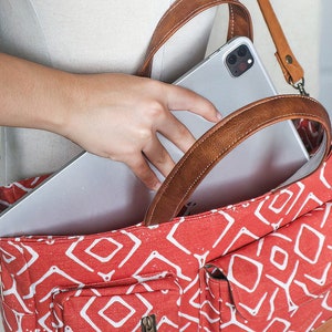 K&ouml;nnte beinhalten: Eine rote und wei&szlig;e gemusterte Canvas-Tragetasche mit braunen Ledergriffen und einer Rei&szlig;verschlusstasche. Ein silbernes Tablet-Computer wird in die Tasche gelegt.