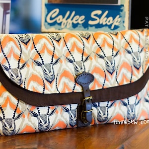 May include: A brown and white fabric clutch bag with a flap closure and a buckle. The bag has a pattern of orange and white antelope heads. The bag has the text "ithinksew.com" printed on the bottom right corner.
