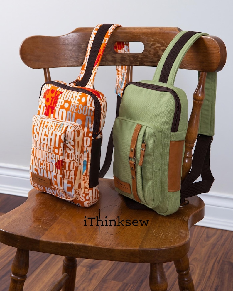 Two handmade Kenzie crossbody sling bags, one in an orange graphic print and one in solid olive green with brown leather accents, resting on a wooden chair.