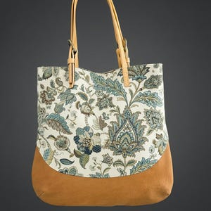 May include: A tote bag with a floral pattern in shades of blue, green, and brown on a cream background. The bottom of the bag is a solid tan leather, and it has tan leather handles.