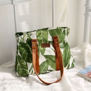 May include: A white canvas tote bag with green leaf print and brown leather straps and handles. The bag has a brown leather tag that says "handmade".