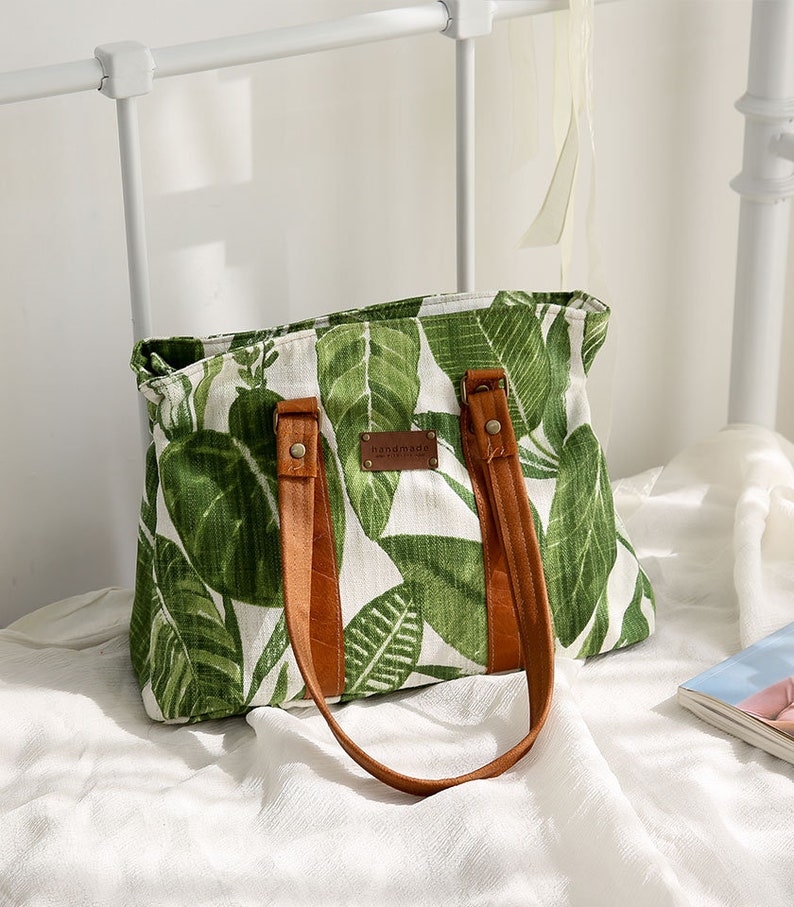 May include: A white canvas tote bag with green leaf print and brown leather straps and handles. The bag has a brown leather tag that says "handmade".