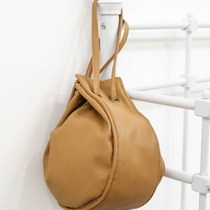 Peut inclure: Un sac seau en cuir marron avec une fermeture &agrave; cordon et deux anses sup&eacute;rieures. Le sac est suspendu &agrave; un support blanc.