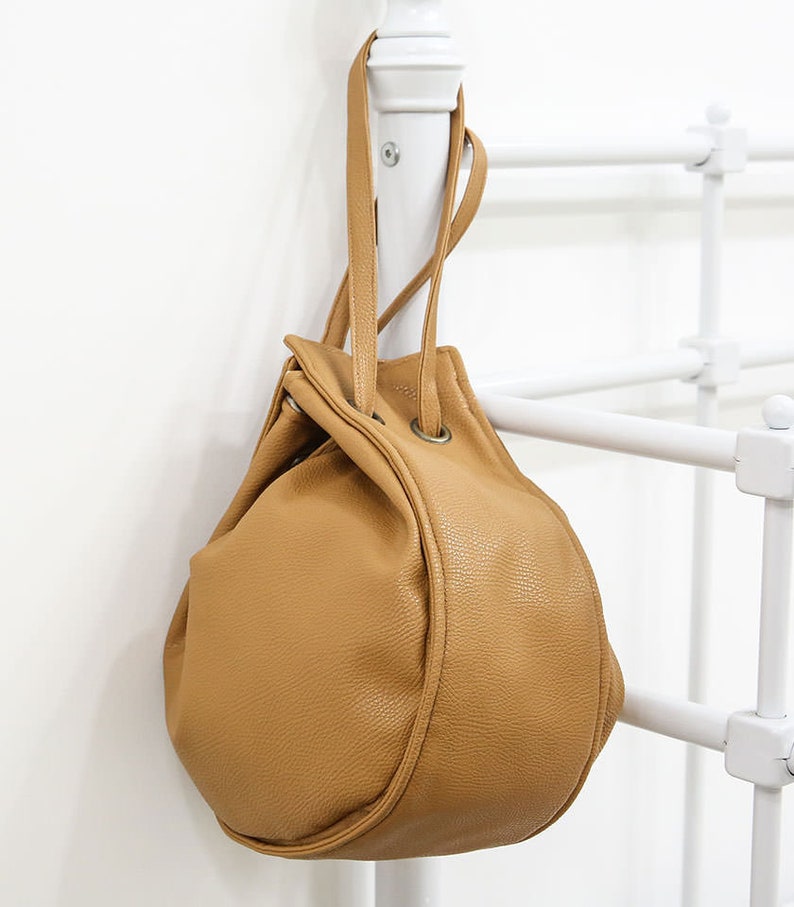 Peut inclure: Un sac seau en cuir marron avec une fermeture &agrave; cordon et deux anses sup&eacute;rieures. Le sac est suspendu &agrave; un support blanc.