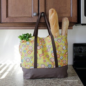 May include: A yellow and brown reusable tote bag with a circle pattern. The bag is filled with a loaf of bread and some green leafy vegetables.