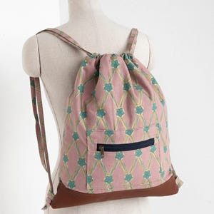 May include: A drawstring backpack featuring a dusty pink fabric with a repeating pattern of teal flowers and yellow crisscross lines. The bag includes a front pocket with a dark blue zip and a solid brown leather-like base with matching straps.
