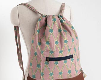 May include: A drawstring backpack featuring a dusty pink fabric with a repeating pattern of teal flowers and yellow crisscross lines. The bag includes a front pocket with a dark blue zipper and a solid brown leather-like base with matching straps.