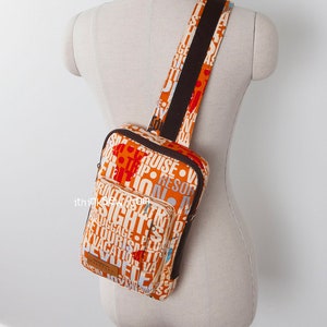 May include: A sling backpack featuring an orange and white fabric with a repeating travel-themed word print including terms like cruise, trip, and tourism. The bag has a brown strap, brown zipper trim, and a small brown rectangular label at the bottom.