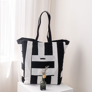 Puede incluir: Una bolsa de mano grande de rayas blancas y negras con un bolsillo más pequeño de rayas blancas y negras en la parte delantera. La bolsa tiene dos asas largas y está sobre una superficie blanca.