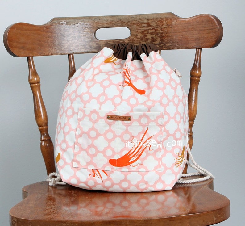 May include: A drawstring backpack featuring a pink and white quatrefoil pattern with orange octopus illustrations. The bag has a brown fabric top closure, a front pocket with a brown leather-like tag labelled HAND MADE, and white rope straps.